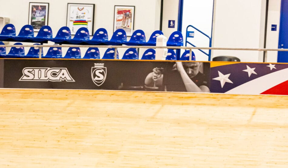A cyclist wearing a red outfit and helmet rides a track bike in an indoor velodrome with wooden flooring. The background features empty blue seats and a large blurred image of another cyclist on the wall above.