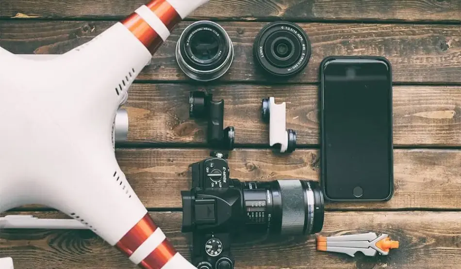 A flat lay photo on a wooden surface at home features a drone, three camera lenses, an action camera, a smartphone, a camera with an attached lens, and a small toy rocket. The items are arranged neatly, suggesting a theme of photography and technology.